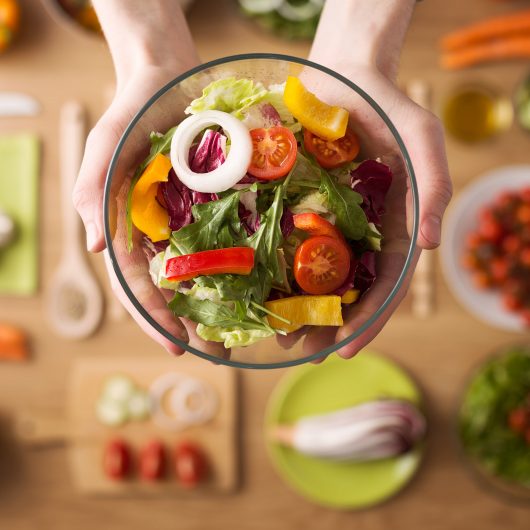 Hands holding an healthy fresh vegetarian salad in a bowl, fresh raw vegetables on background, top view